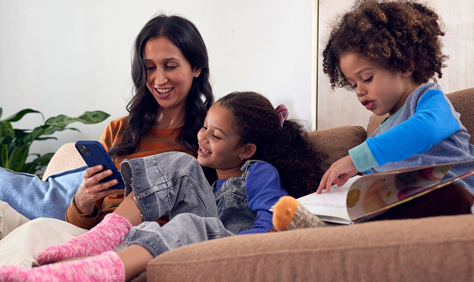 A family sits on a couch.  A mother and child are looking at their phone while another child reads a book next to them.