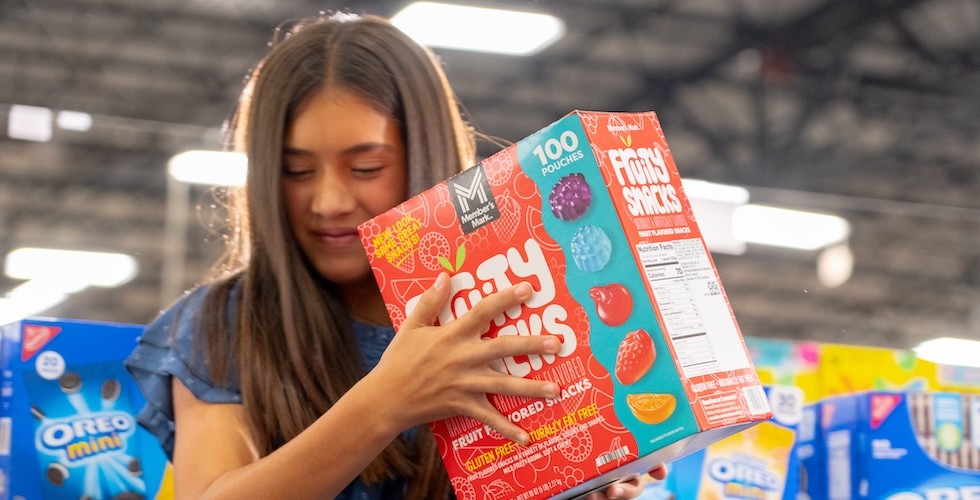A young girl smiles while packing fruit snacks into a shopping cart inside of a Sam's Club.