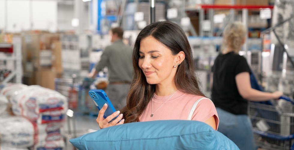 A Sam's Club member smiles while inspecting a pillow and checking its features using her phone.