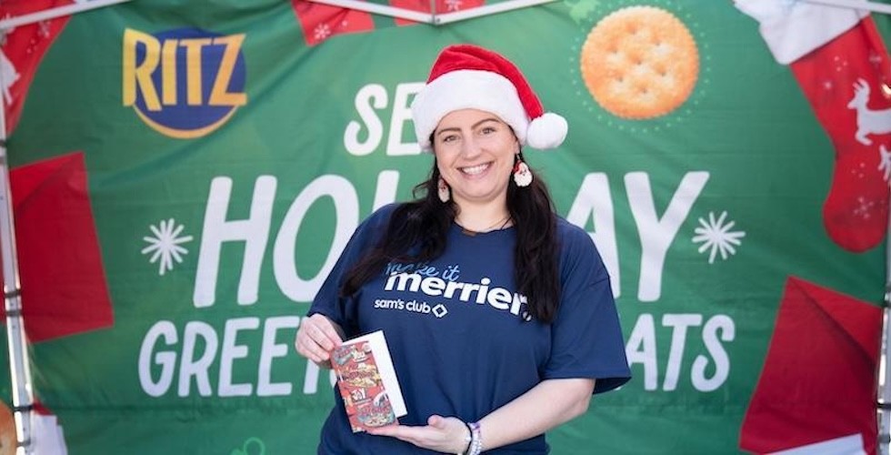 A woman wearing a Santa hat holds a card, smiling warmly at the camera.