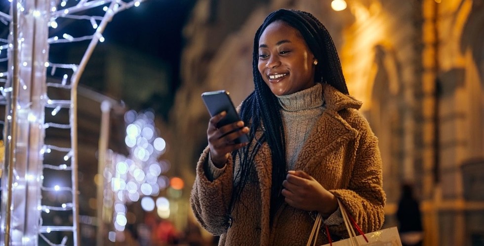 A woman using her phone while walking in a brightly lit city street at night.